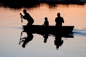 A fisherman stands on a boat with fishing gear, surrounded by calm waters and greenery on Norfolk Broads.