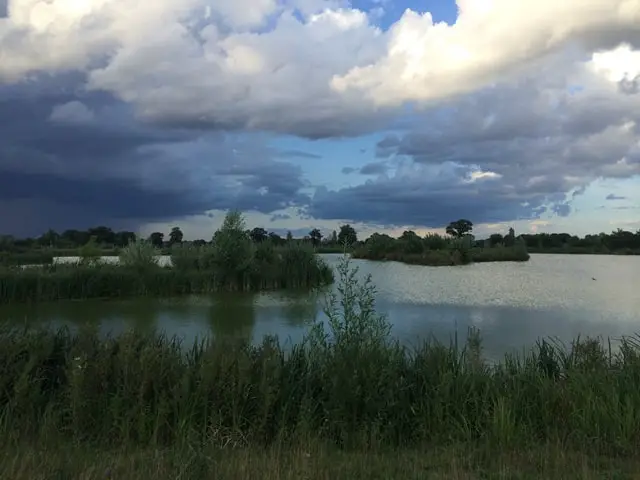 View of Airfield Lakes in Norfolk. A peaceful lake surrounded by greenery with a wooden platform and a fishing rod in the foreground