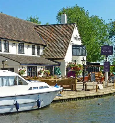 A view of a restaurant on the Norfolk Broads. The picture shows a large wooden deck extending over the water, with several tables and chairs set up for dining. The restaurant building is visible in the background, with large windows overlooking the water. The sky is clear and blue, and boats can be seen passing by on the water.