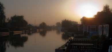 View of River Bure in Norfolk. A serene river surrounded by trees, with boats and a bridge in the distance.