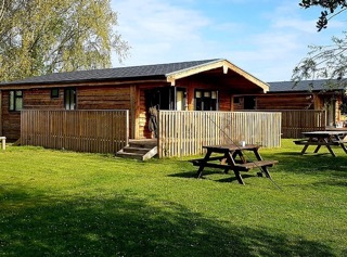 Top 5 fishing lakes with accommodation in Norfolk An idyllic view of Woodlakes, Norfolk, showcasing tranquil waters surrounded by lush greenery, with charming wooden cabins nestled among the trees.