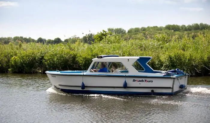 An image of a leisurely picnic boat on a Norfolk broads Wroxham trip gliding smoothly on calm waters. The boat is adorned with a colorful picnic setup, complete with a checkered blanket, a wicker basket filled with goodies, and comfortable seating. Surrounding greenery and a serene ambiance complete the idyllic setting for a relaxing day out on the water.